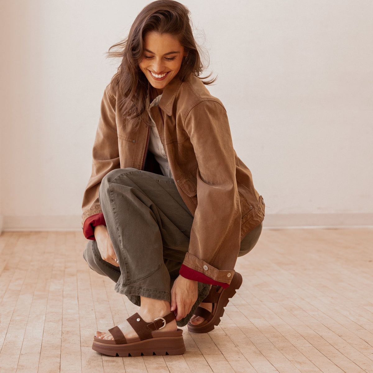 Young woman crouching indoors wearing Vista Luxe Liah facing left, brown suede platform sandals with adjustable back strap and arch support
