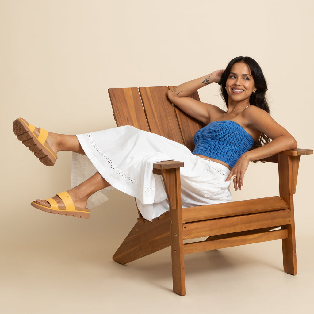 Woman seated in wooden chair wearing Vista Luxe Thea sandals facing left, showcasing lightweight golden apricot sandals with arch support.