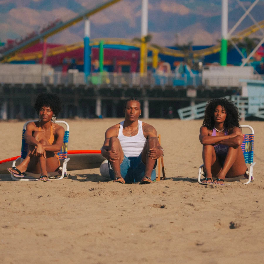 Three adults sitting on beach chairs wearing REEF X Ebony Beach Club Original Stripes sandals facing forward on sand.