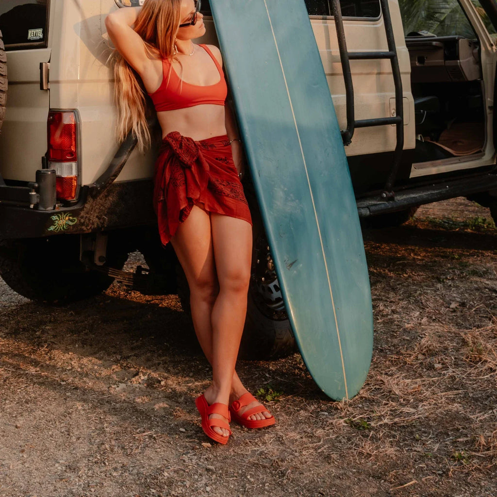 Woman wearing red Vista Capri sandals facing forward, standing beside a van with a surfboard in a beach setting.